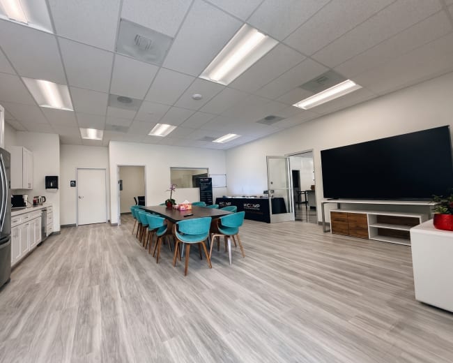 The image shows a modern office kitchen area with a dining table, chairs, a refrigerator, and a large television.