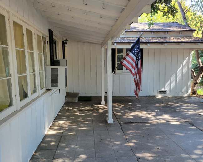 The image shows a white house exterior with a porch featuring a flagpole holding an American flag, on a sunny day with dappled shadows on the concrete floor.
