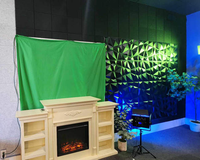 A green screen is set up in front of a decorative fireplace, flanked by textured black and gray walls with ambient lighting.