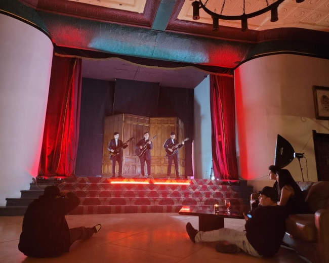 Three musicians perform on a stage with red lighting, while an audience sits on the floor and a couch in a well-lit venue.