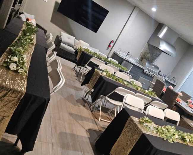 A spacious indoor event area features multiple tables covered with black tablecloths and adorned with gold sequin runners, surrounded by white chairs, and a large television mounted on the wall.