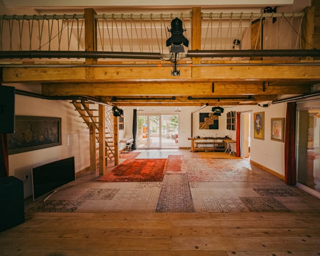The image shows a spacious interior with wooden beams, a staircase, and area rugs on the floor, leading to large glass doors that open to an outdoor area.