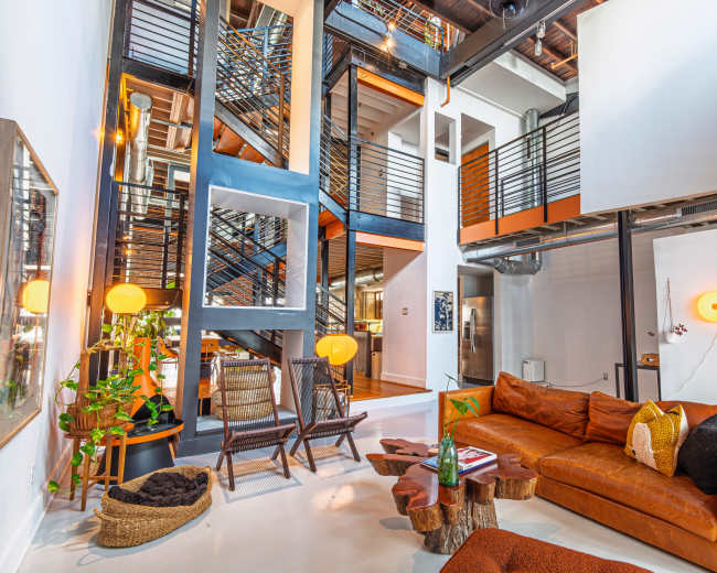 The image shows a modern, multi-level living space featuring a brown leather sectional sofa, a wooden coffee table, and a staircase with metal railings.