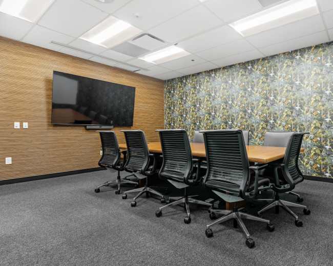 The image shows a modern conference room with a large wooden table surrounded by black swivel chairs, a flat-screen TV mounted on a wooden wall, and a floral wallpaper accent.