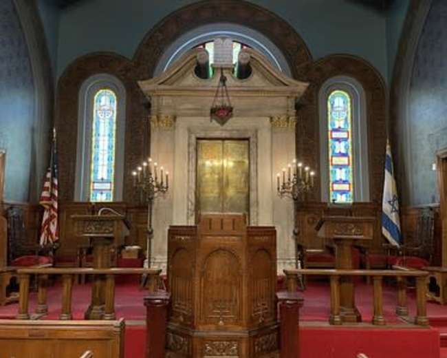 The interior of a synagogue features a pulpit in the foreground, flanked by wooden chairs, with ornate stained glass windows and a large central ark behind it.