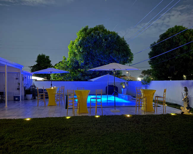 The image shows a backyard with a swimming pool illuminated by blue lighting, surrounded by yellow bar tables and umbrellas under a night sky.