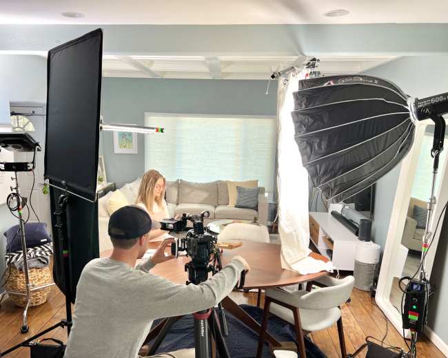A person sits at a table in a living room while a crew sets up video lighting and camera equipment around them.