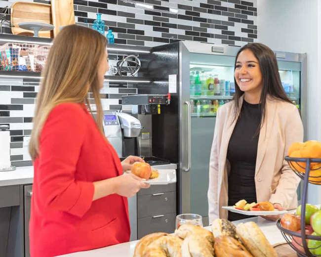 Two women are engaged in conversation near a food display in a modern kitchen with a black and white tiled backsplash.
