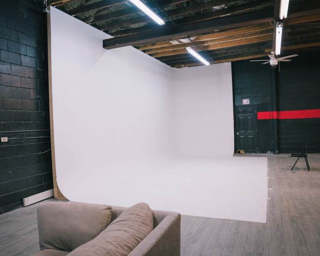 The image shows a spacious studio with a white curved backdrop, wooden beams on the ceiling, and a gray sofa in the foreground.