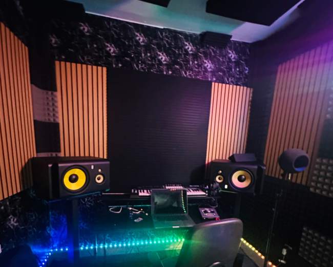 The image shows a music studio featuring a desk with audio equipment, two speaker monitors, and colorful LED lights along the floor.