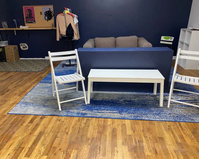 The image shows a room with a brown couch against a blue wall, two white chairs, and a light-colored table on a blue rug.