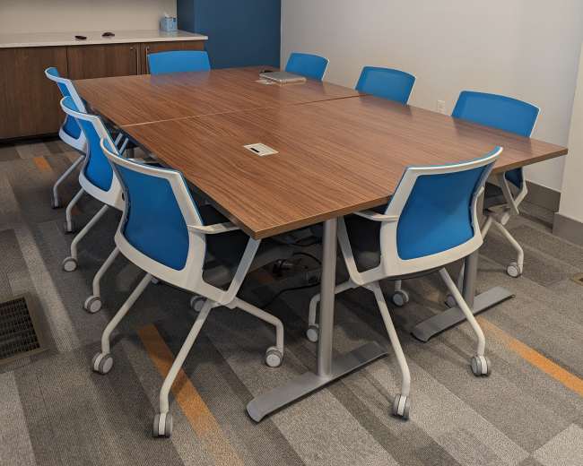 A modern conference room features a large brown table surrounded by eight blue and white rolling chairs, with a wall-mounted screen in the background.