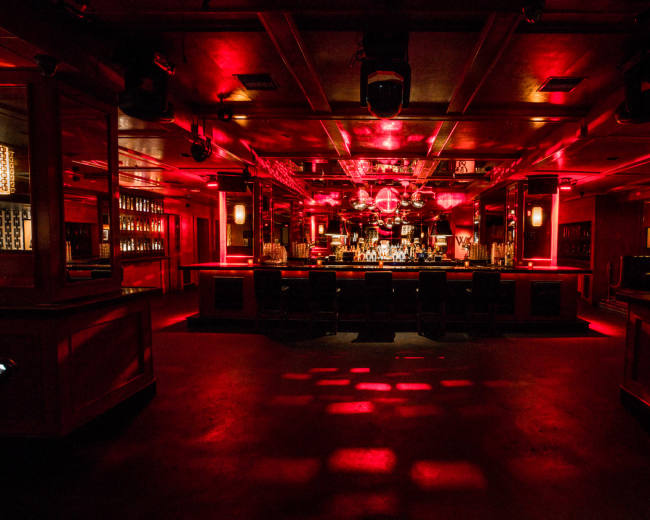 The image shows a dimly lit bar with red lighting, a central mirrored disco ball, and a long bar counter lined with stools.