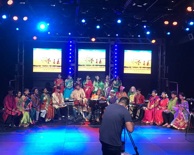 A diverse group of musicians and singers dressed in colorful traditional attire performs on stage with instruments and lighting.