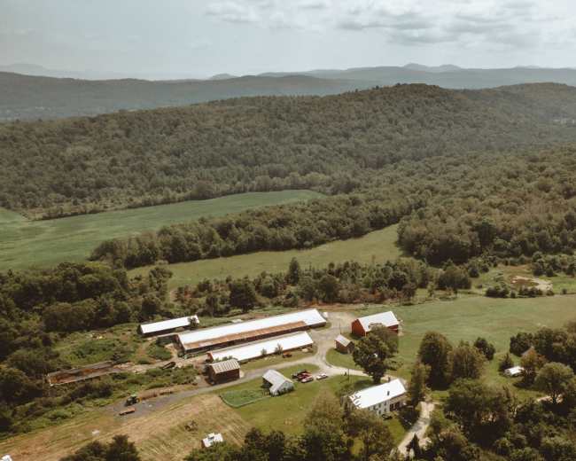 A rural landscape with a farm nestled among rolling hills and dense forests.