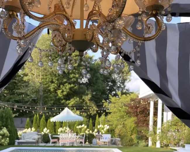 A spacious backyard featuring a swimming pool surrounded by lush greenery and outdoor furniture, with a chandelier hanging in the foreground.