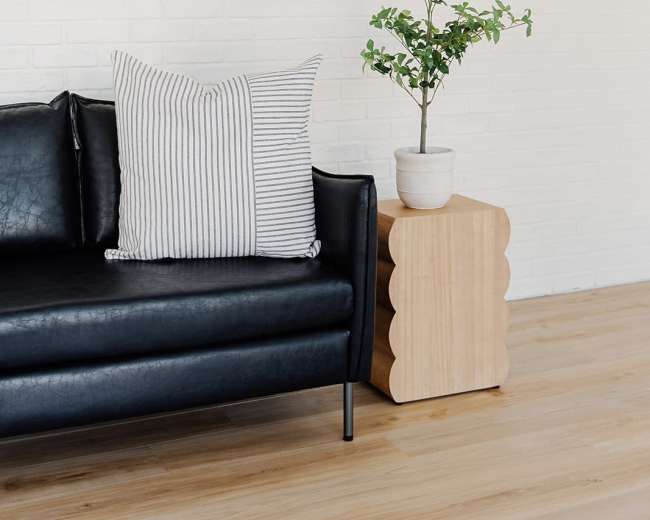 A black sofa with striped pillows is positioned next to a wooden side table that holds a potted plant against a white brick wall.