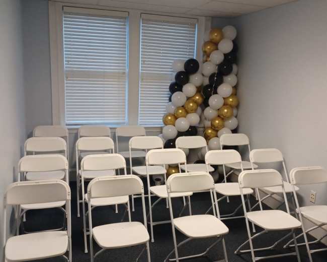 A small room is set up with rows of white folding chairs facing a cluster of black, white, and gold balloons.