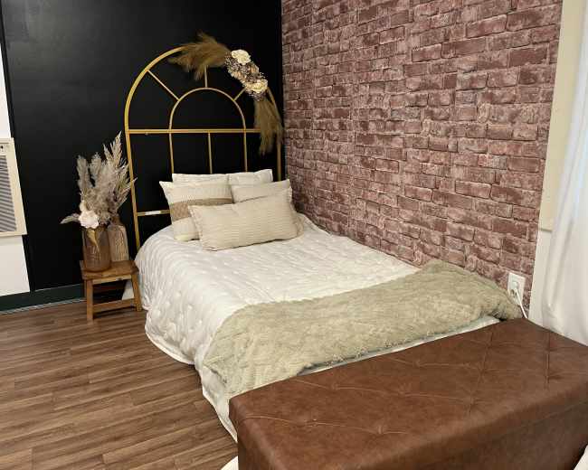 The image shows a cozy bedroom with a bed featuring a gold arched headboard, layered with white and beige bedding, against a brick-patterned wall.