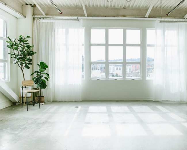 A spacious room features large windows covered with sheer curtains, a potted plant, and a wooden chair against a light-colored wall.
