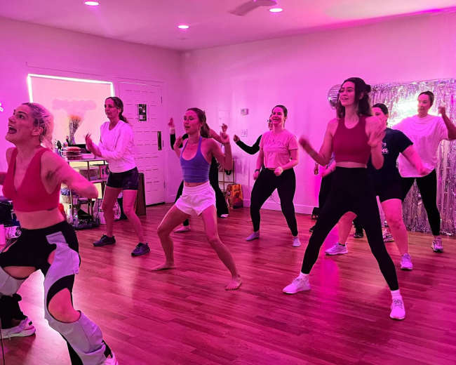A group of eight individuals is participating in a dance class in a brightly lit, pink-themed studio with a mirror.