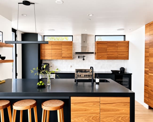 The image shows a modern kitchen with wooden cabinetry, a black countertop, and a minimalist design featuring a large island with bar stools.