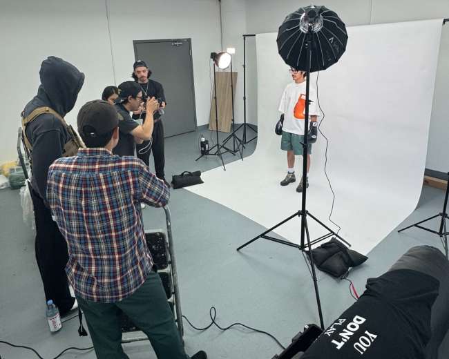 A group of five individuals is gathered in a studio, preparing for a photoshoot with a model in front of a white backdrop and lighting equipment.