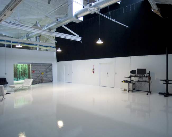 The image shows an empty studio space with a bright, reflective white floor, black curtains, and a workstation against one wall.