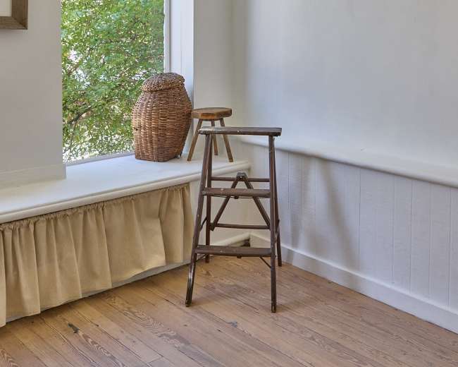 The image shows a wooden stool placed next to a window with a view of green foliage outside.