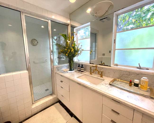 The bathroom features a glass shower enclosure, a marble countertop with a sink, modern fixtures, and large windows that let in natural light.