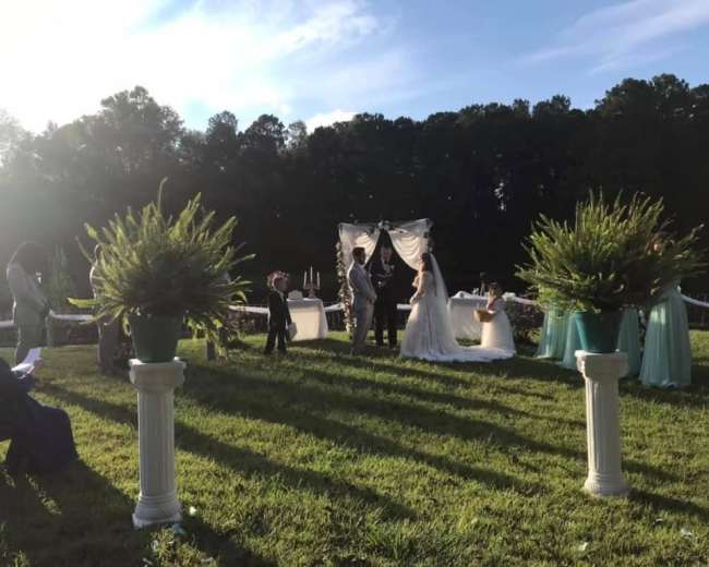 A couple stands under a decorated arch while exchanging vows in an outdoor wedding ceremony surrounded by guests.