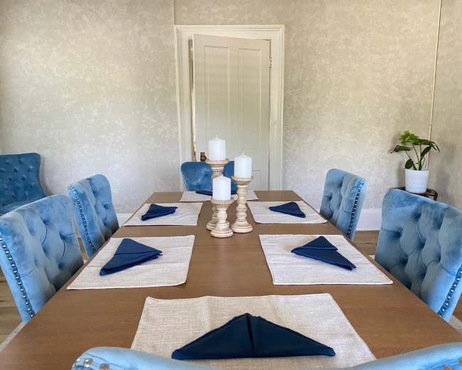 A dining table is set with beige placemats and blue napkins, surrounded by light blue upholstered chairs, with candles at the center and a potted plant in the background.