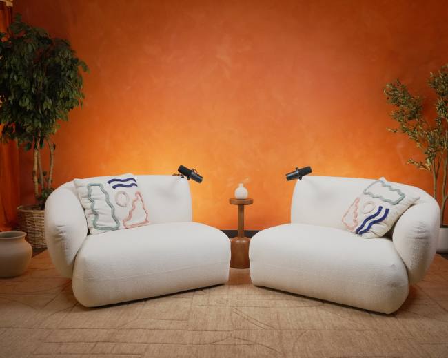 The image shows two white, plush chairs facing each other, with a small round table and decorative plants against an orange wall.
