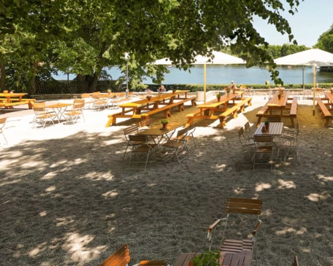 A sandy outdoor seating area with wooden tables and chairs under umbrellas, adjacent to a calm body of water and surrounded by trees.