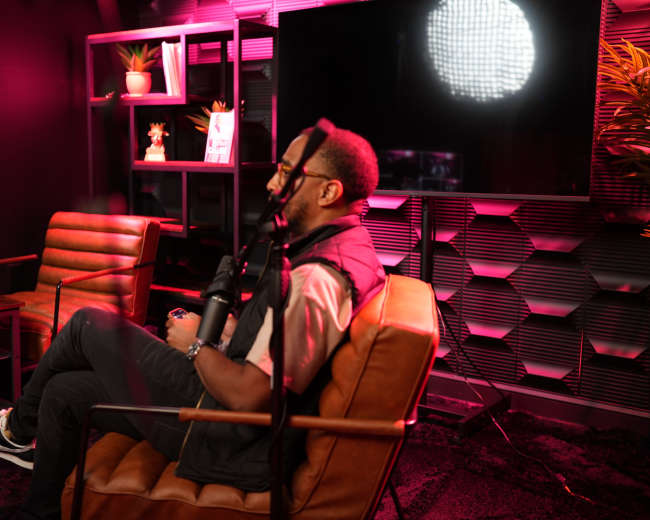 A person sits in a chair with a microphone in front of them, surrounded by pink lighting and modern decor.