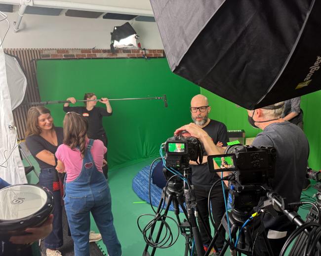 A group of people is in a studio equipped with green screens and various cameras, preparing for a shoot.