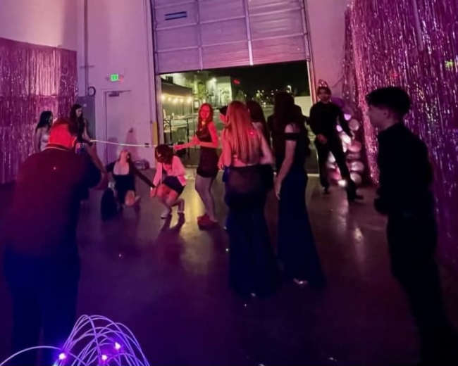 A group of people is participating in a limbo game at a party, with a shiny, pink backdrop and various costumes present.