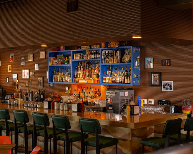 The image shows a bar with a wooden counter, several high chairs, and a colorful backlit shelf displaying various bottles of liquor and framed photos on the walls.