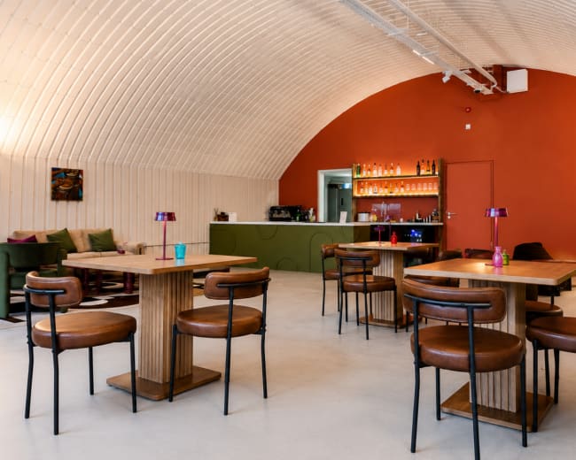 The image features a modern café interior with wooden tables and chairs, a bar area in the background, and leafy plants decorating the space.
