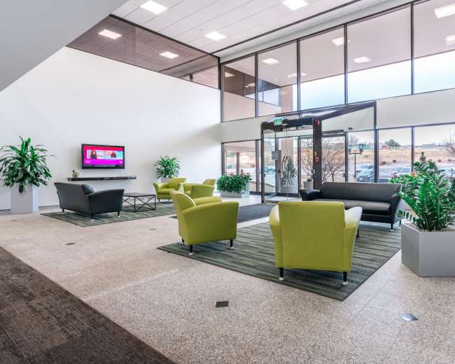 A modern lobby area with green chairs, a dark sofa, television, and large windows allowing natural light to enter.
