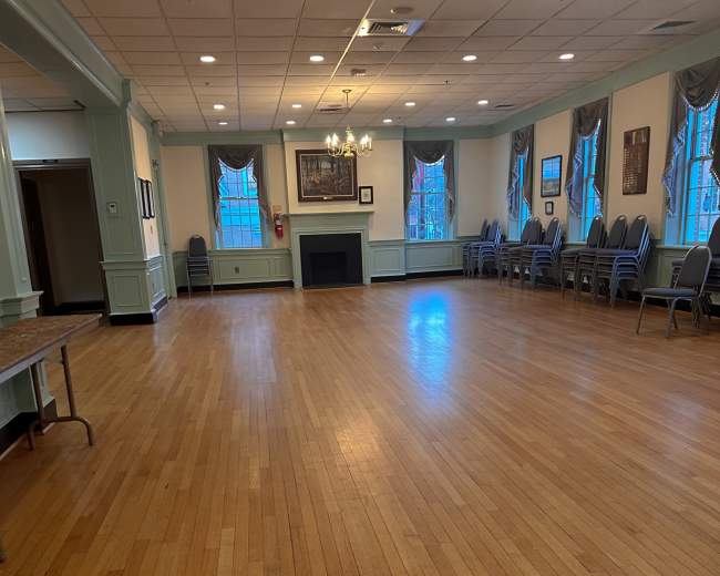 A spacious, empty hall with wooden floors, bright overhead lights, and windows lined with drapes.