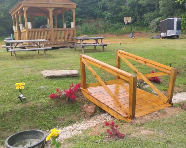 A wooden bridge spans a small flower bed with red and yellow flowers, leading to a gazebo in a grassy area surrounded by trees and picnic tables.