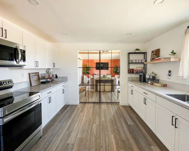 A modern kitchen features white cabinetry, stainless steel appliances, and a clear glass partition leading to a living area with plants and a television.