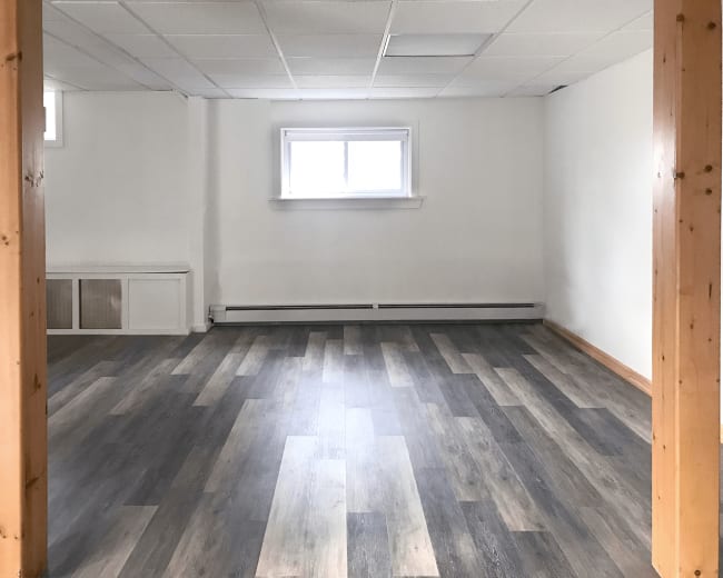 A spacious room with wooden beams, light-colored walls, a window, and a floor featuring gray and brown vinyl planks.