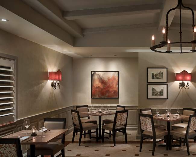The image depicts a warmly lit restaurant interior featuring wooden tables and chairs, along with artwork on the walls and soft lighting from wall sconces.
