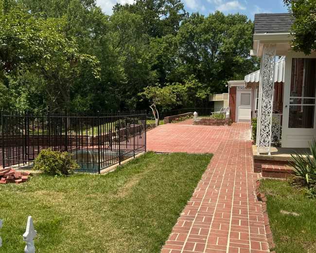 A brick pathway leads through a green lawn alongside a fenced pool area and a house with white features under a blue sky.