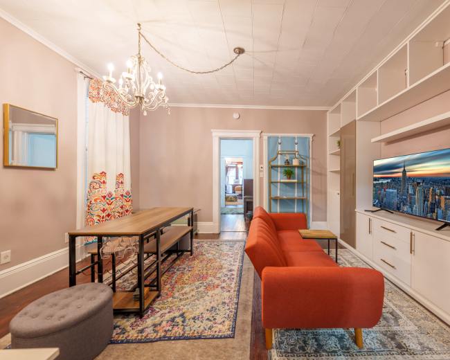 A living room with a red sofa, a dining table, a television stand, and decorative elements including a chandelier and patterned curtains.