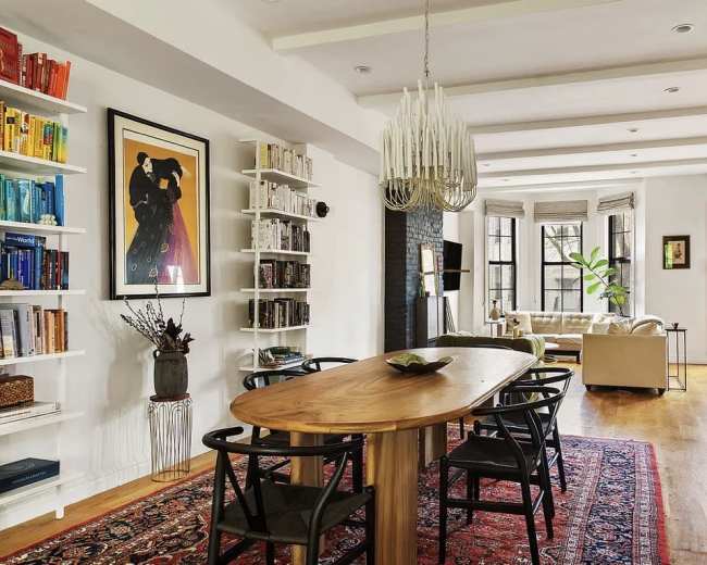 The image shows a spacious, modern living and dining area with a large wooden dining table, bookshelves filled with books, and a cozy seating arrangement.