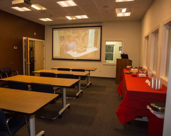 A conference room is set up with several tables and chairs facing a projected screen, alongside a food table covered with a red cloth.