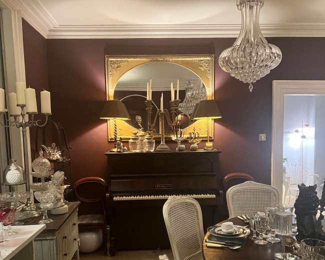 A wooden piano is positioned against a wall with a large mirror and candelabra, surrounded by dining tables set with glassware and silverware in a warmly lit dining room.